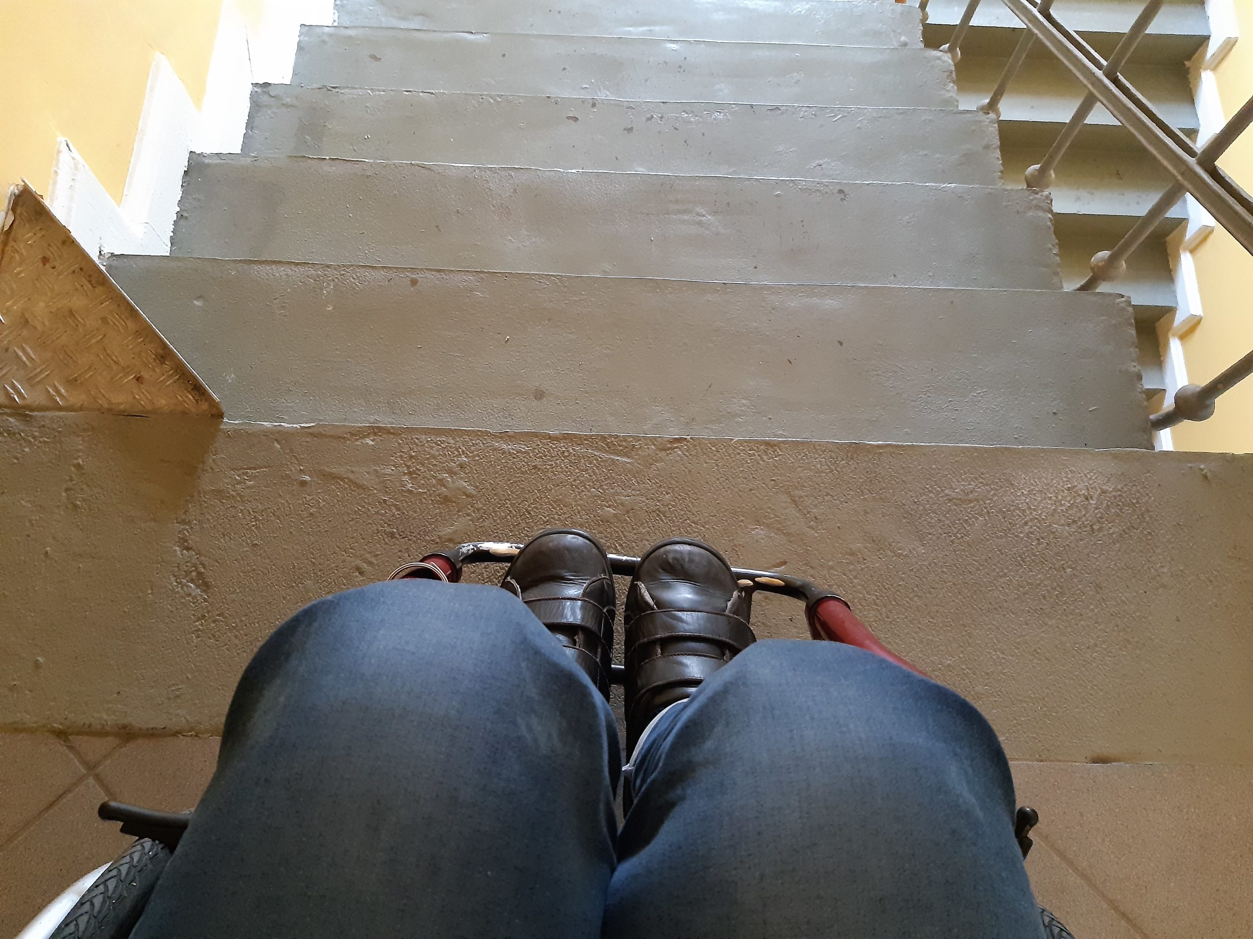 Unterkörper einer Rollstuhl fahrenden Person aus Vogelperspektive vor Treppe